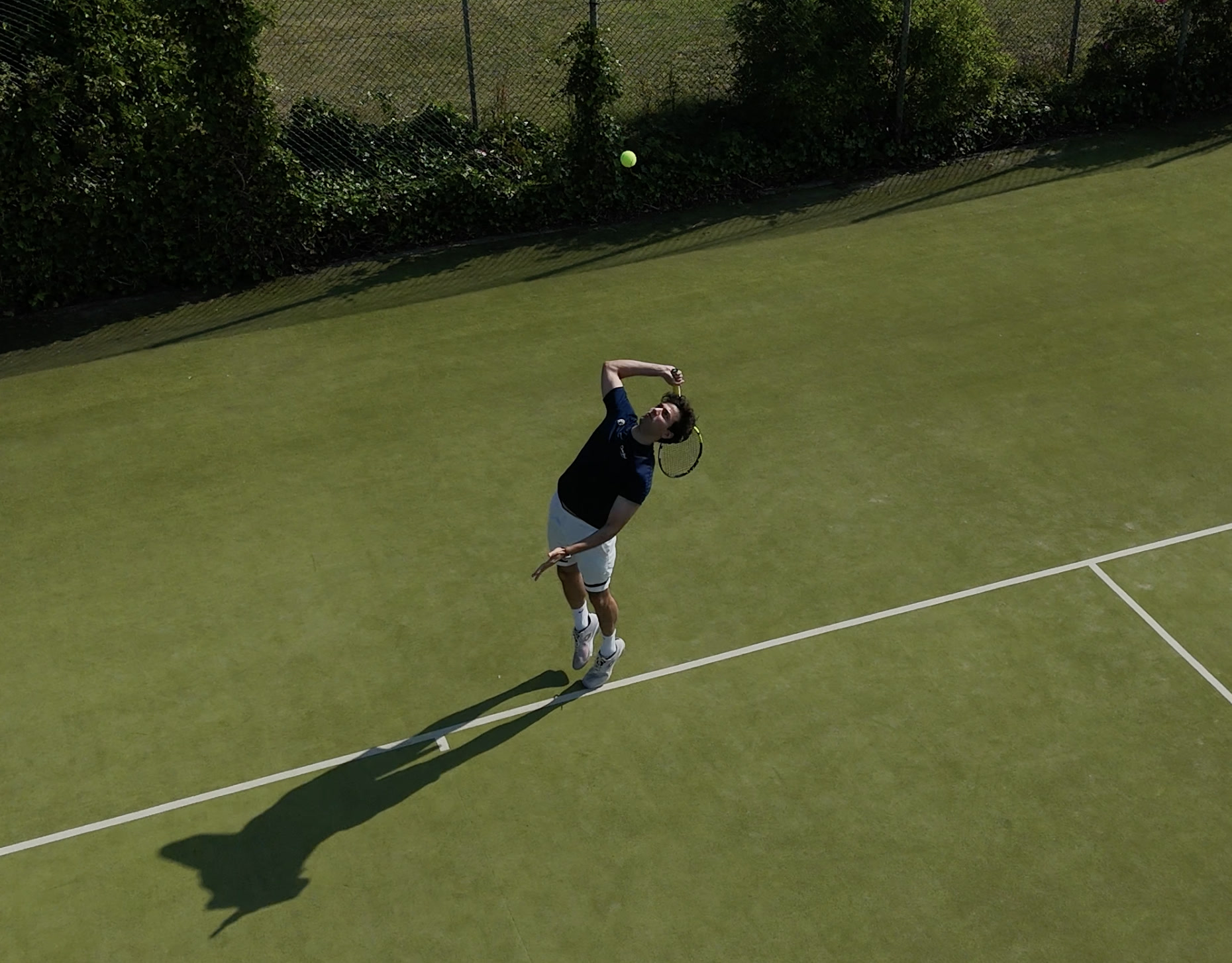 Drone shot of a tennis serve on a grass court, ball tossed high above the player