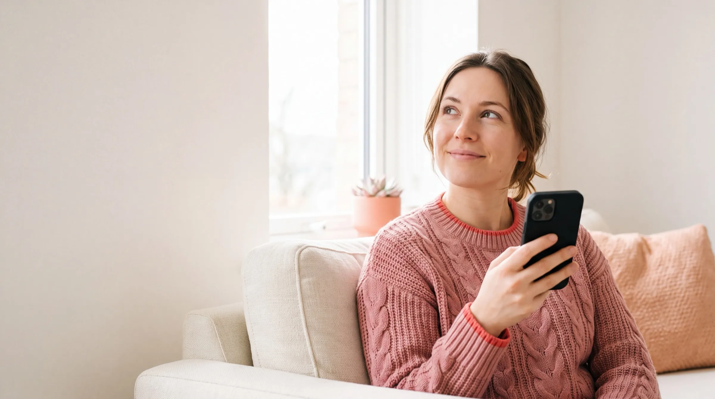 A young woman at home on a sofa glances up from her phone with a relaxed smile